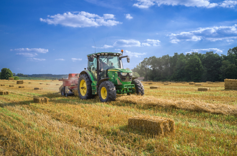 A tractor harvesting hay from a large field. A bale of hay in the foreground.